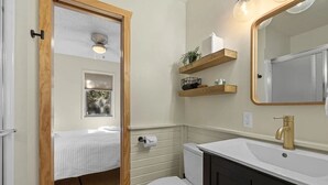 Casa de banho compacta com sanita, lavatório e espelho. Existem prateleiras de madeira sobre a sanita e uma cama visível através de uma porta aberta.