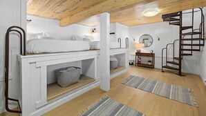 Quarto pequeno e bem iluminado com beliche, uma escrivaninha, uma escada e um espelho redondo na parede.