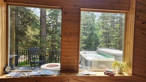 Uma cabana de madeira com grandes janelas dando para uma floresta. No interior, há uma mesa com uma cadeira, um livro e uma pequena planta.