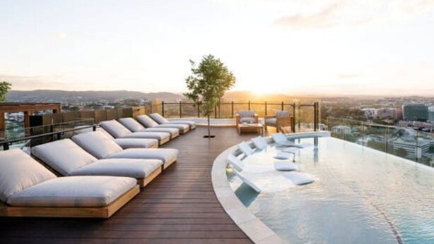 Une piscine à débordement située sur le toit, avec des chaises longues et une terrasse en bois, offrant une vue imprenable sur le paysage urbain au coucher du soleil.