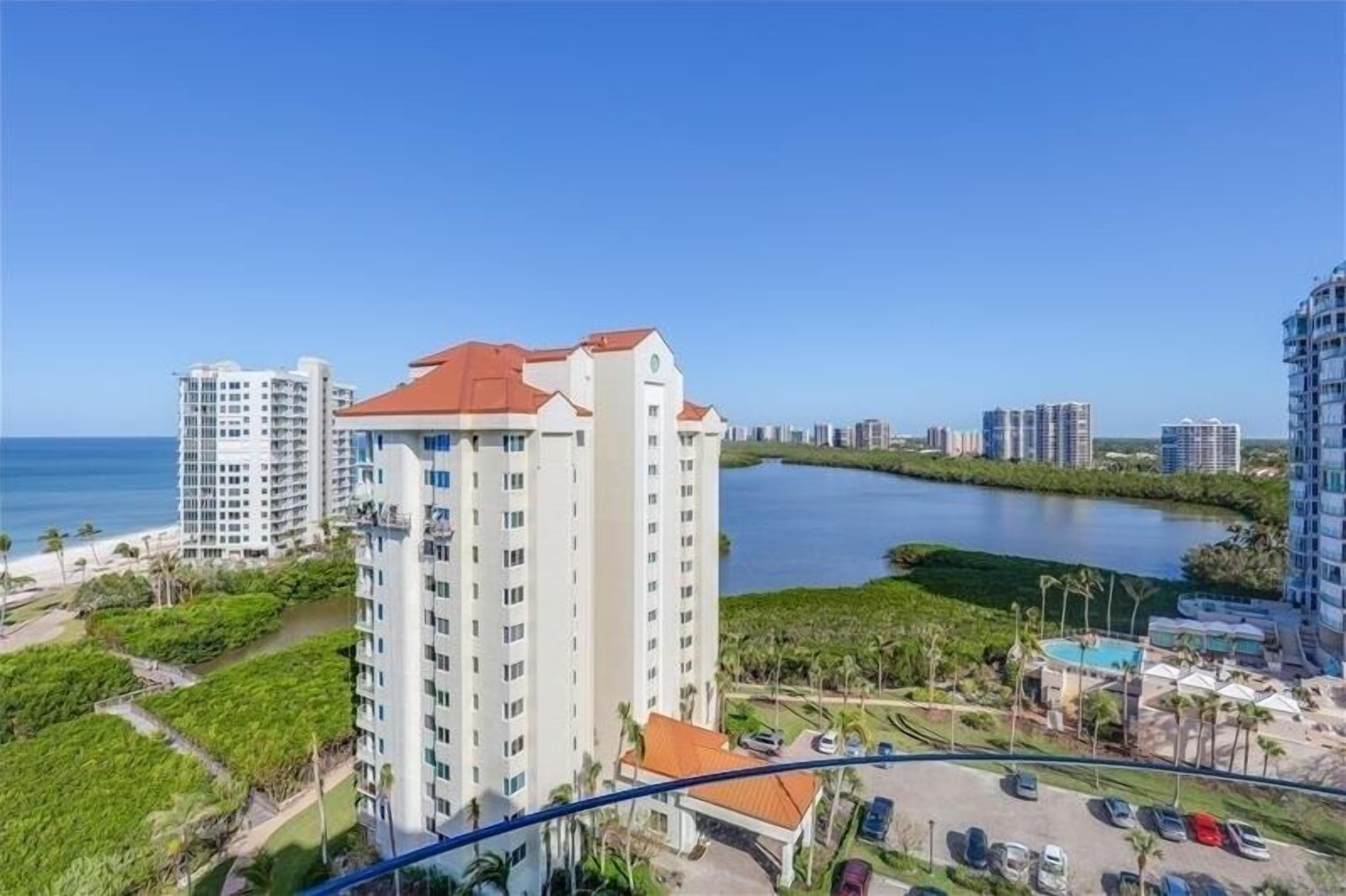 A high-rise hotel with a red roof, surrounded by greenery and a body of water.