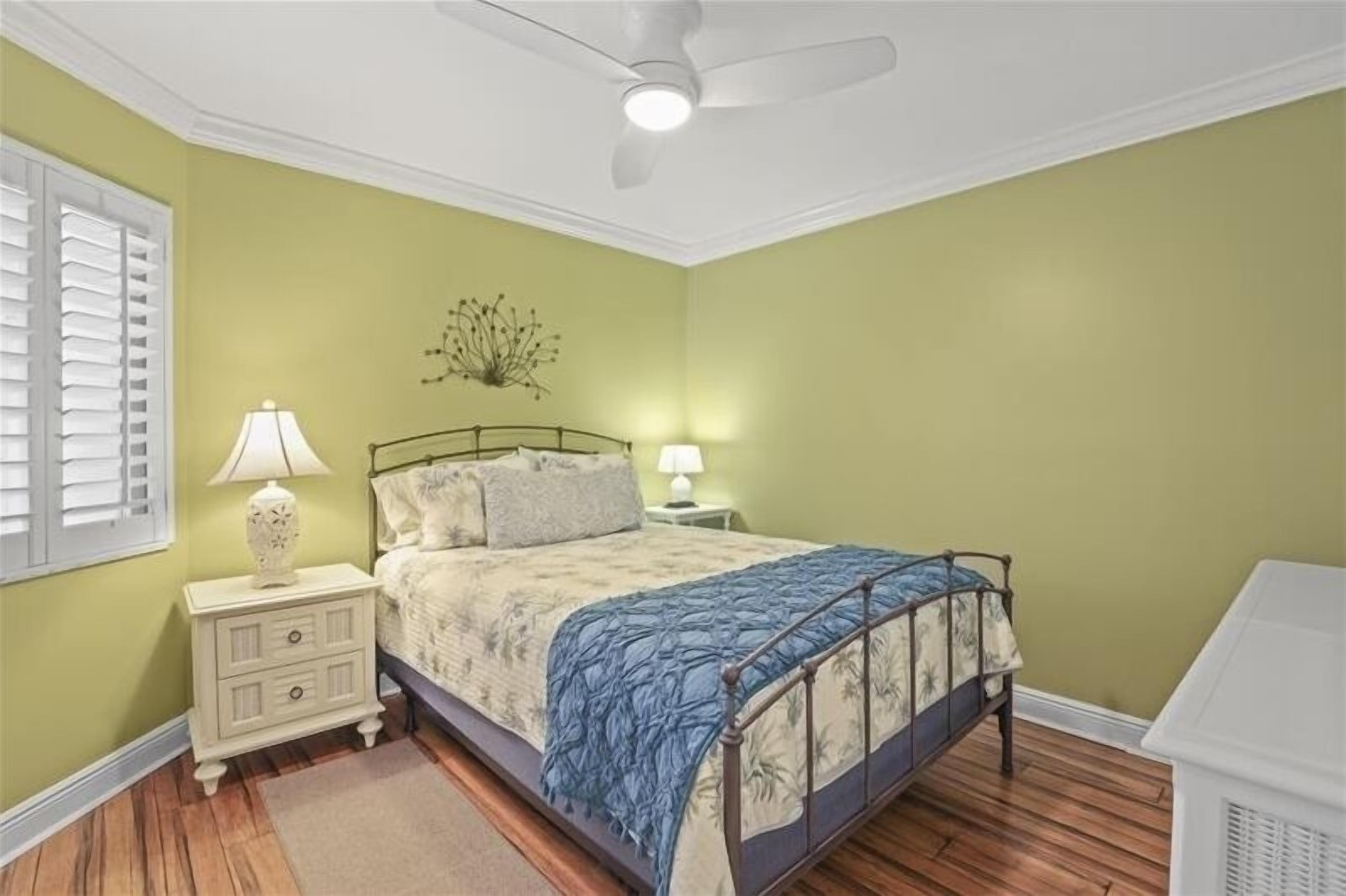 A bedroom with a bed, nightstands, lamps, a ceiling fan, and a window with shutters.
