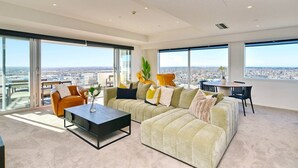 Un salon moderne avec un canapé d’angle, une table basse et un coin repas comprenant une table et des chaises. De grandes fenêtres offrent une vue sur la ville.