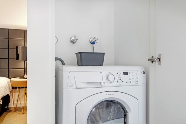 Une petite buanderie moderne et fonctionnelle, équipée d’un lave-linge à chargement frontal, d’un étendoir et d’un petit évier.
