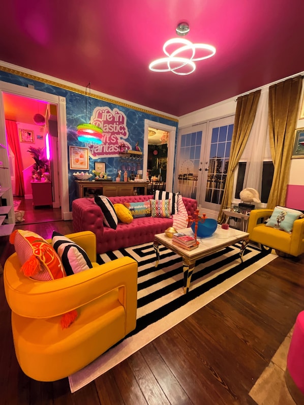 A brightly lit living room with a pink sofa, yellow armchair, and a striped rug.