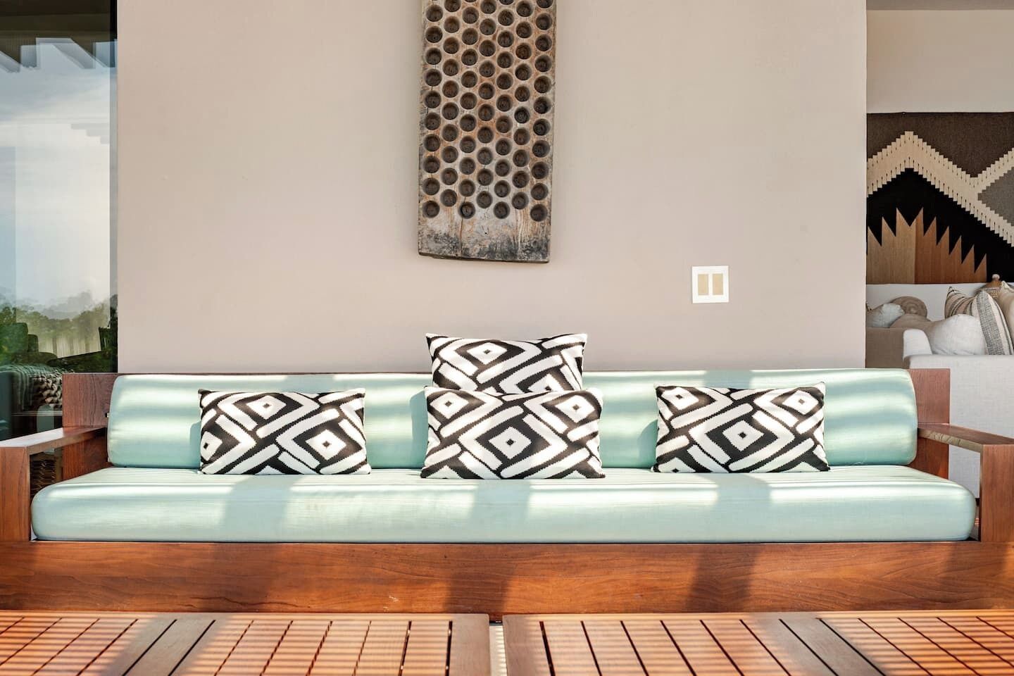 Un'area salotto esterna moderna con un divano azzurro chiaro e cuscini decorati, pavimento in legno e un elemento decorativo a parete.