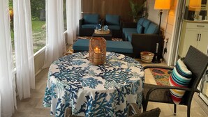 Un rincón al aire libre acogedor con una mesa cubierta con un mantel estampado en azul y blanco, rodeado de sillas de mimbre y un sofá con cojines azules.