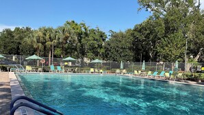 Una piscina con agua azul cristalina, rodeada de sillas de descanso y sombrillas, y una cerca con árboles al fondo.