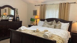 Una cama bien hecha con sábanas blancas, una lámpara de noche y un espejo sobre un tocador.