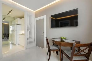 A modern dining area with a round table, two wooden chairs, a flat-screen TV mounted on the wall, and a kitchen visible through an open door. - ORCHID SUITES (Palm Beach)