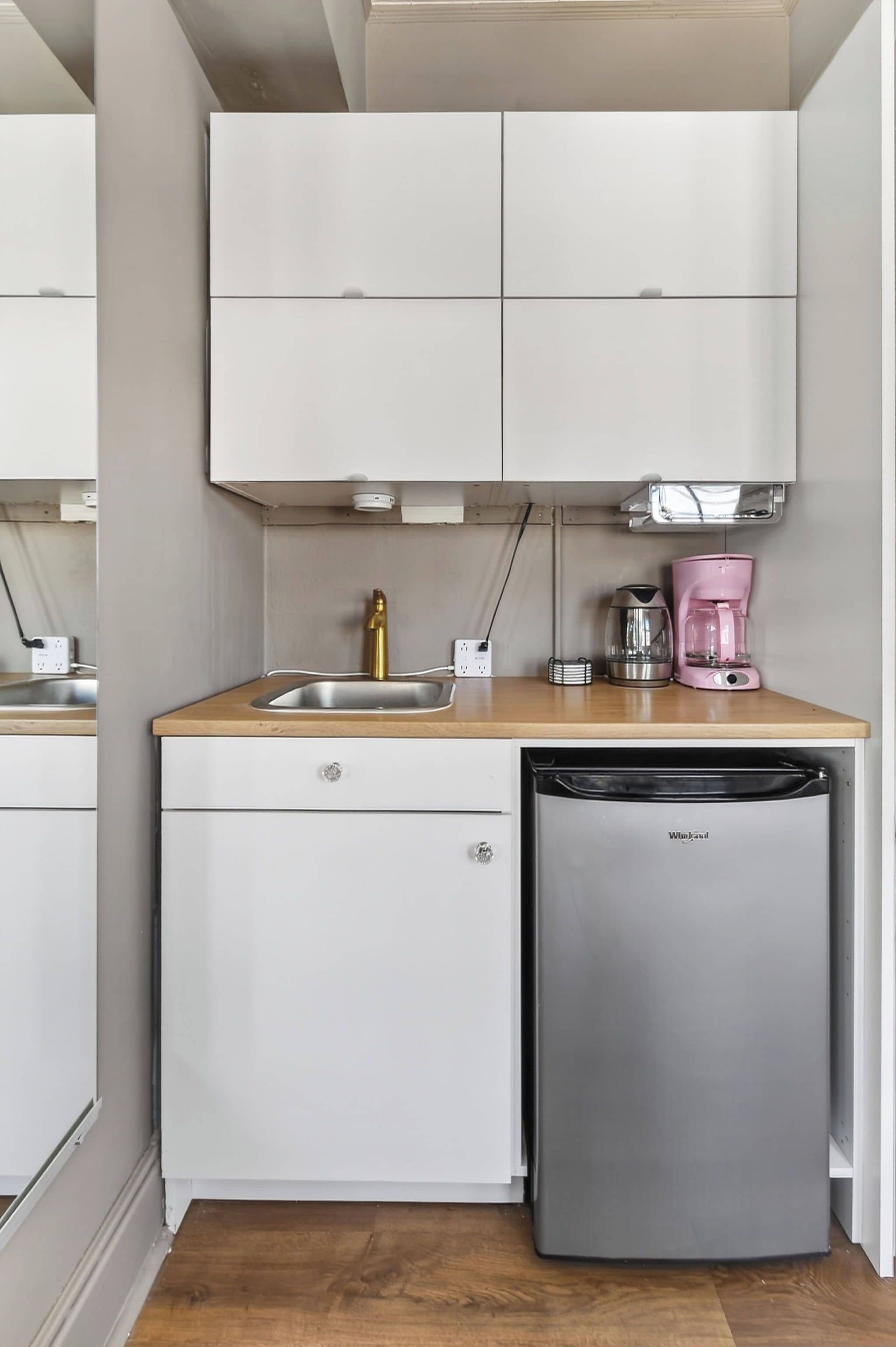 Een compacte keuken met een houten aanrecht, een roestvrijstalen spoelbak, een gouden kraan, een roze koffiezetapparaat, een zilveren waterkoker en een inbouwvaatwasser.