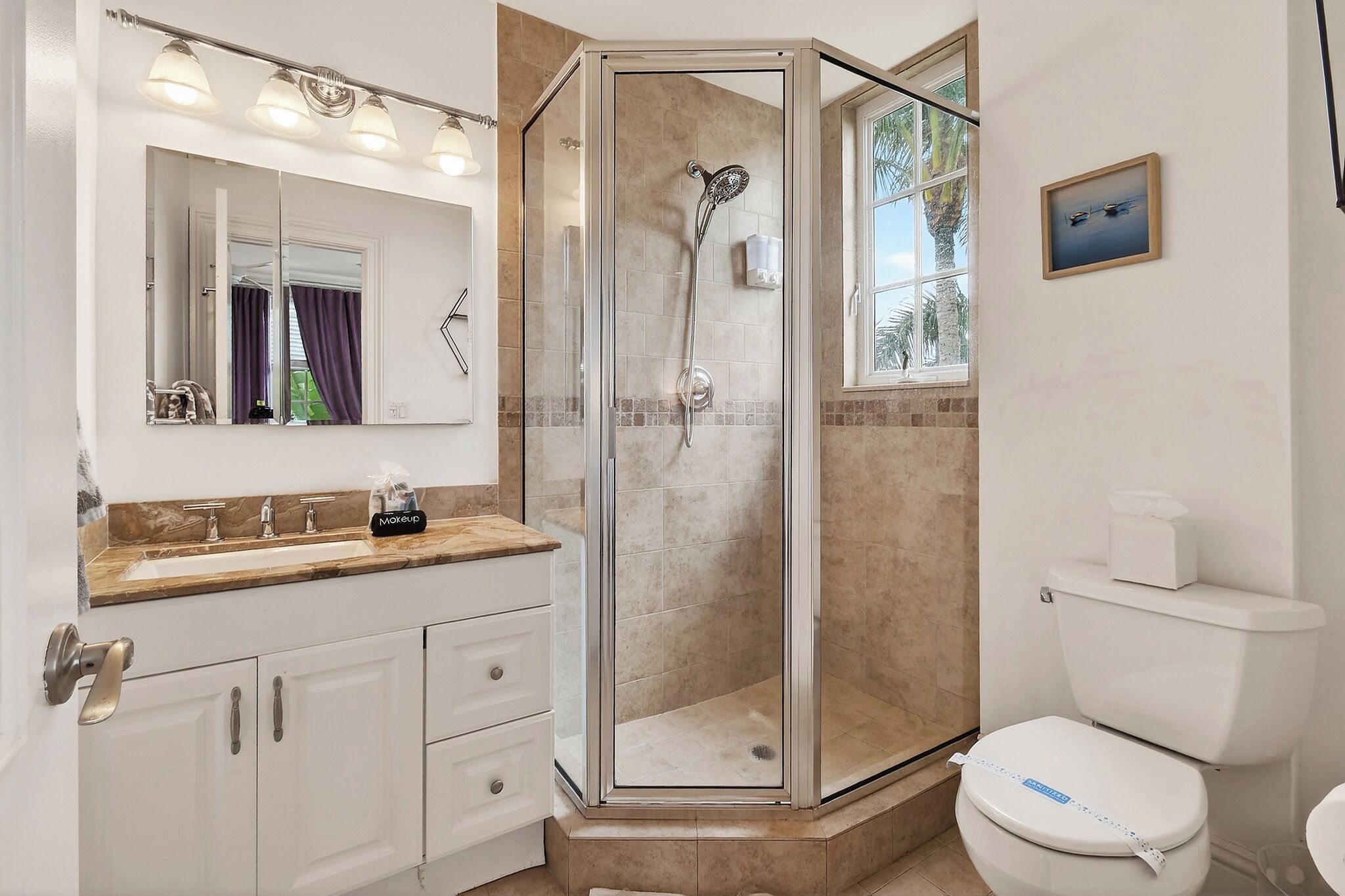 A bathroom with a glass-enclosed shower, a white toilet, a vanity with a mirror, and a window with a view of palm trees.