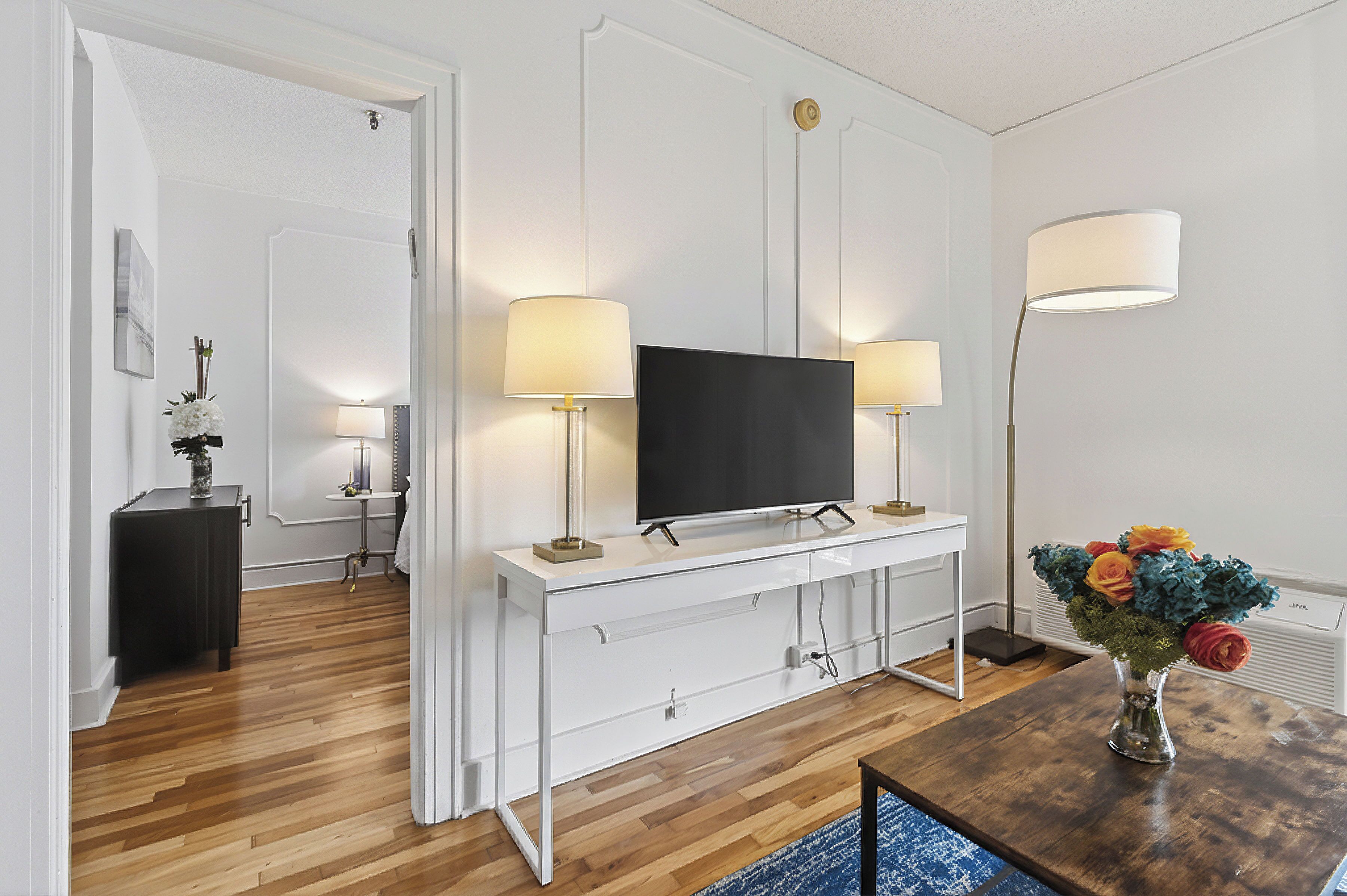 A modern living room with a flat-screen TV, white console, two lamps, a vase with flowers, and a wooden floor.