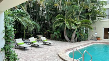 Zona junto a la piscina con sillones de descanso y una pequeña mesa, rodeada de exuberante follaje tropical.