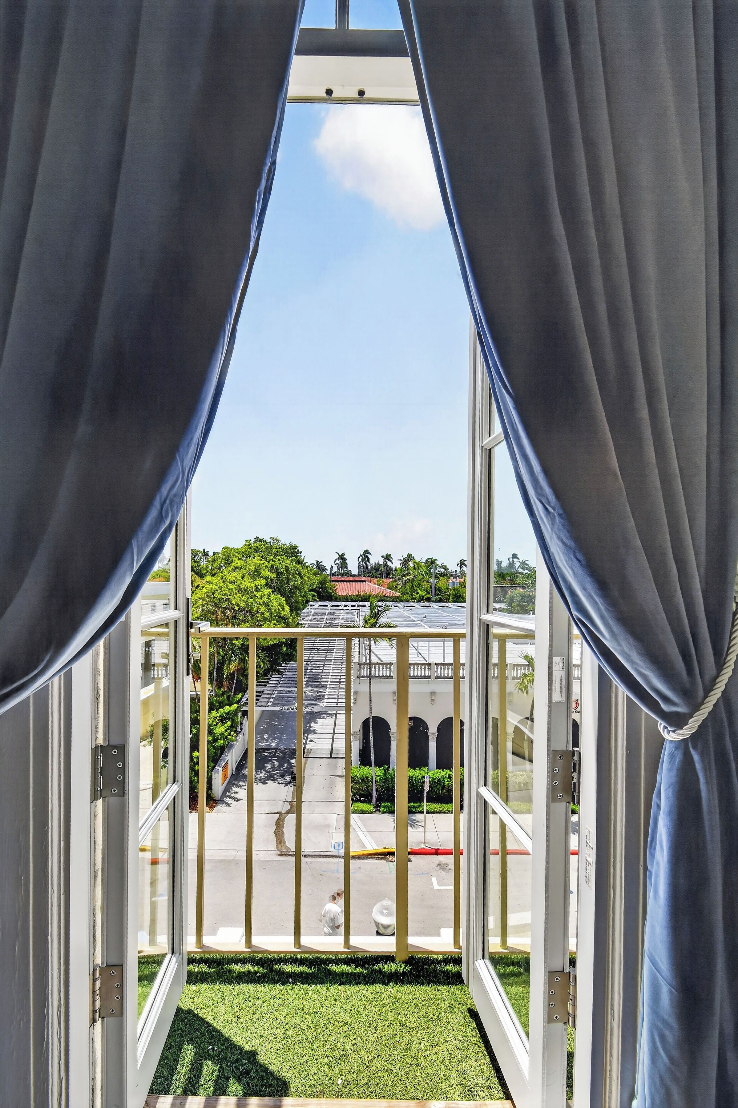 An open window with curtains, a view of a building with solar panels, and a green lawn.