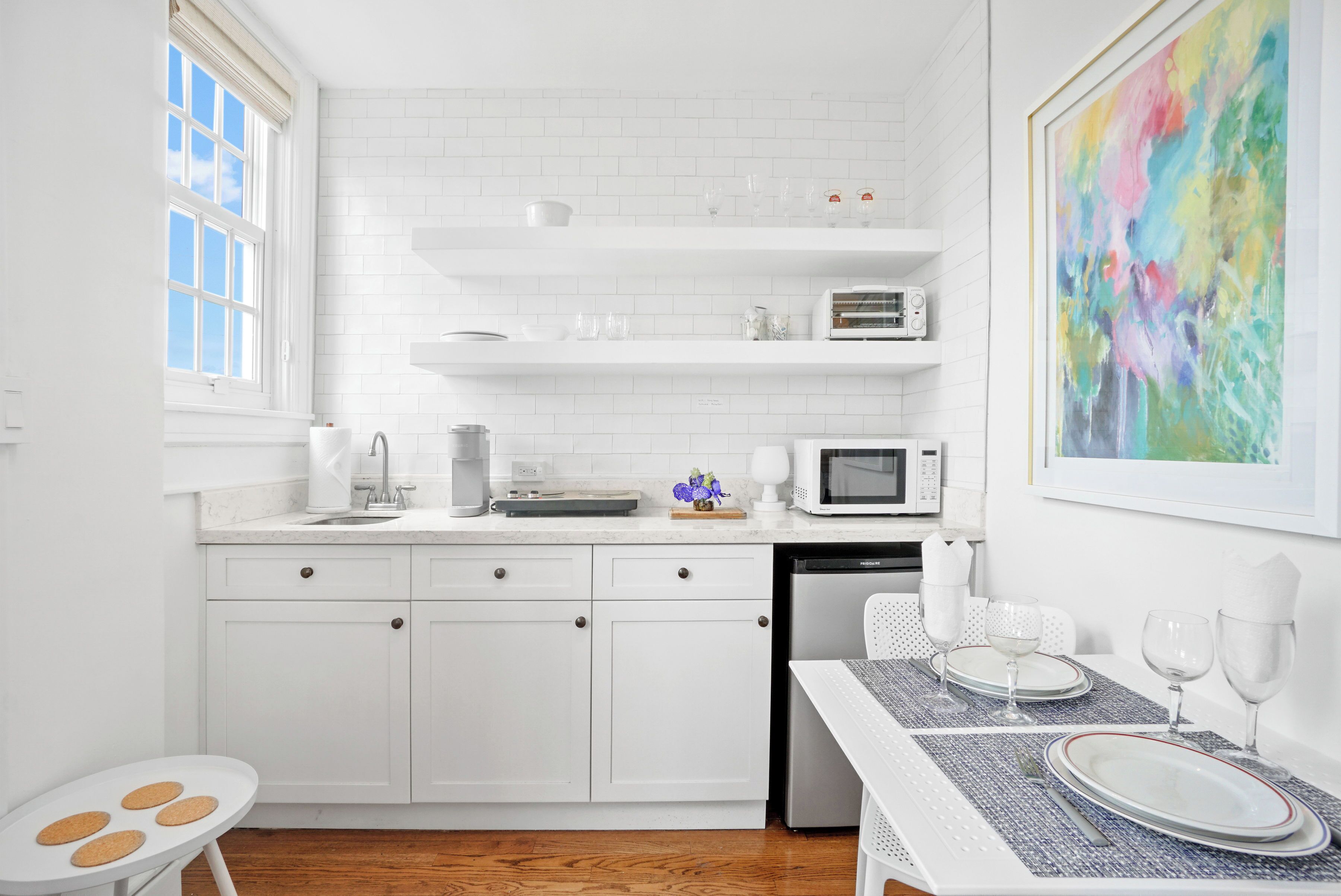 Una cocina moderna con armarios blancos, una zona de comedor con mesa para dos, y un cuadro abstracto colorido en la pared.