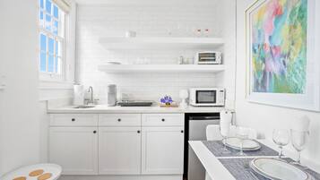 Una cocina moderna con armarios blancos, una zona de comedor con mesa para dos, y un cuadro abstracto colorido en la pared.