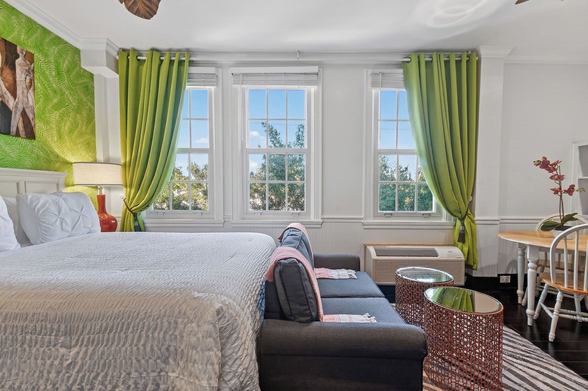 Quarto de hotel com cama, sofá, mesa e cadeira. Há janelas com cortinas, um quadro na parede e um abajur.