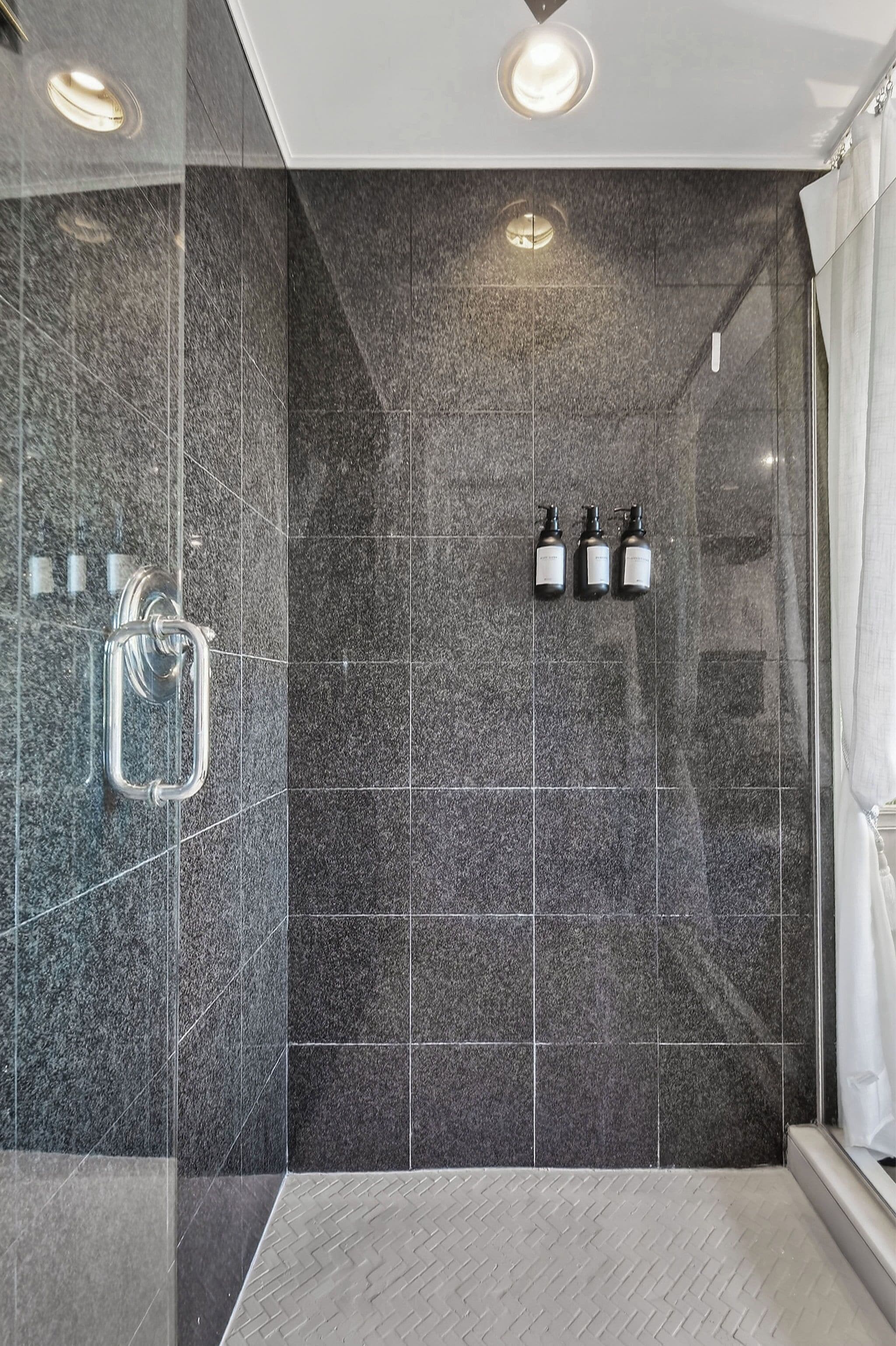 A modern shower area with dark tiles, a glass door, and three bottles of shampoo.