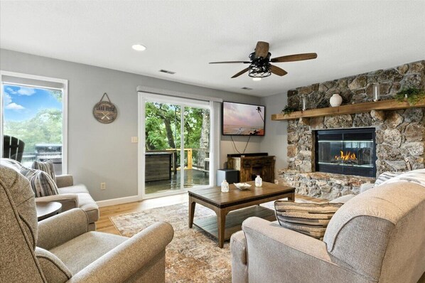 A living room with a stone fireplace, a ceiling fan, and a large TV.