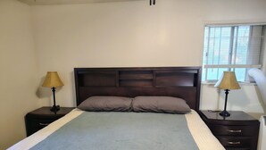 Uma cama com cabeceira, duas mesas de cabeceira com abajures, uma janela com persianas e um ar-condicionado fixado na parede.