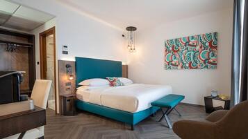 A modern hotel room with a teal bed, a desk, a chair, a bedside table, and a wall-mounted artwork.