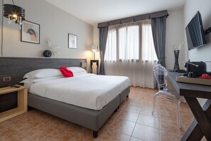 A hotel room with a bed, a desk with a chair, a television, and a window with curtains.