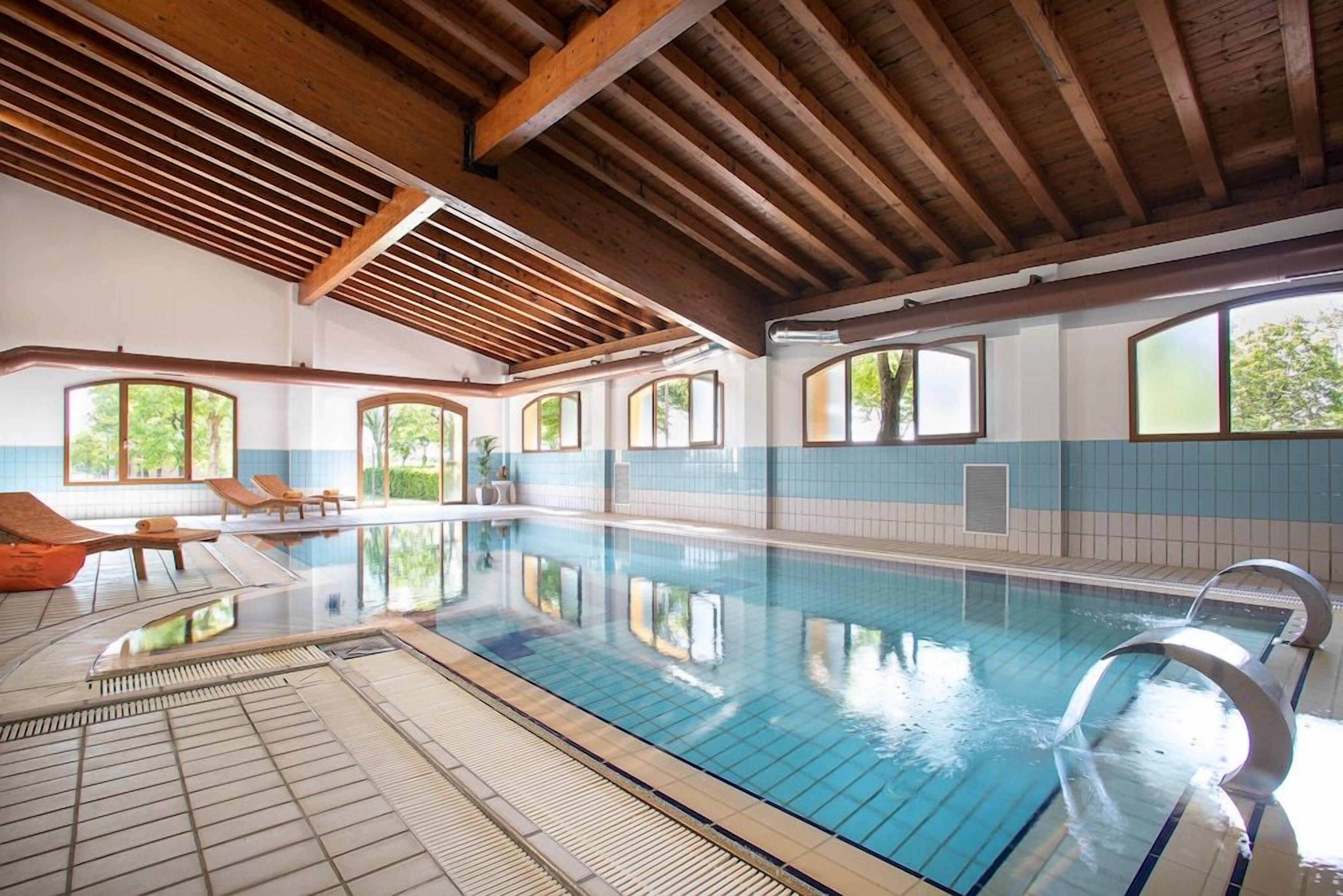 An indoor swimming pool with clear water, wooden beams on the ceiling, and windows with views of greenery.