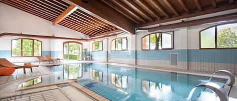 An indoor swimming pool with clear water, wooden beams on the ceiling, and windows with views of greenery.