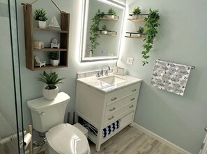 Un baño con un lavamanos blanco, un espejo, un inodoro y estantes con plantas.