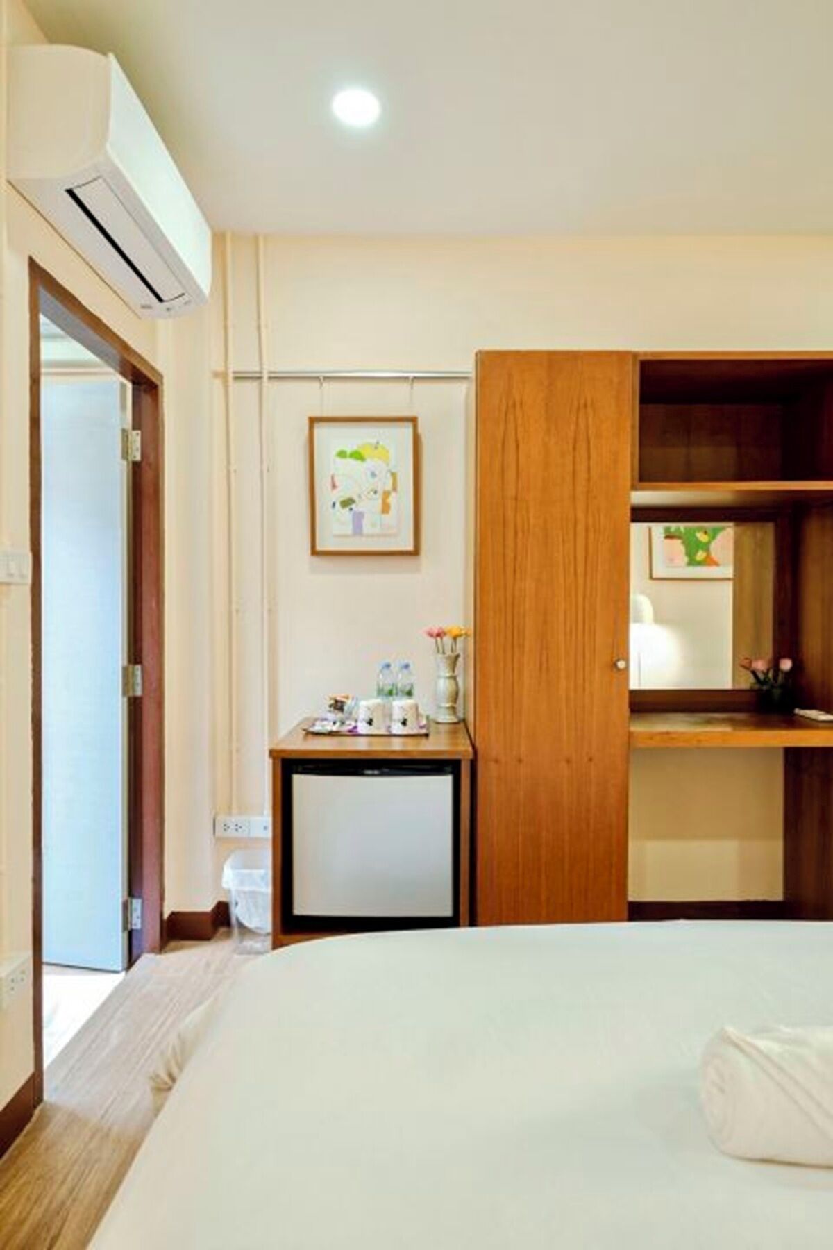A hotel room with a bed, a wooden desk with a vase of flowers, a framed picture on the wall, and an air conditioning unit.