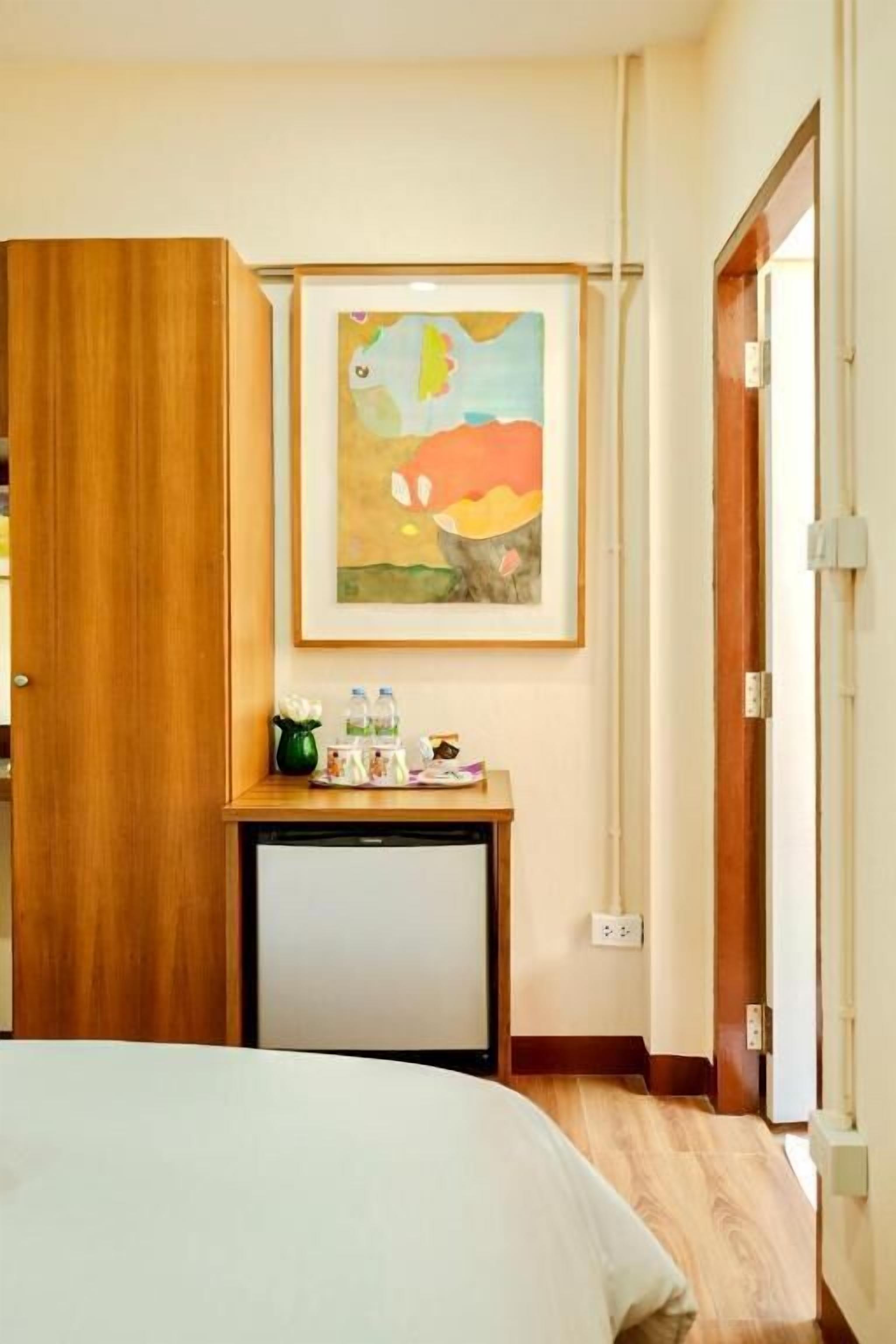 A hotel room with a bed, a small desk with a plant, a framed painting, and a wooden wardrobe.