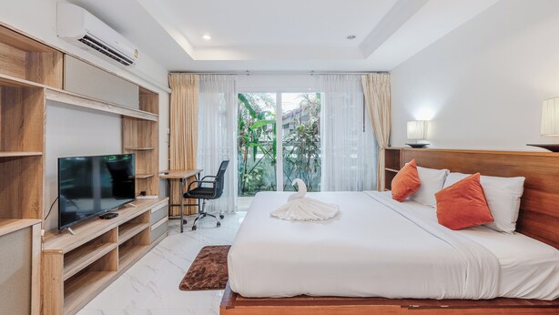 A hotel room with a large bed, a TV on a wooden stand, a desk, and a view of greenery outside. - 1BR Near Naiharn Pool&Relaxed Vibes C199 (Rawai)