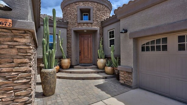 A house with a stone facade, a wooden door, and a garage. There are large cacti in pots in front of the house.