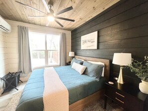 A bedroom with a bed, a ceiling fan, a nightstand with a lamp, a window with curtains, and a framed picture on the wall.