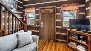A log cabin interior with a wooden staircase, a gray sofa, a television on a wooden stand, and a door with a sign. - 21 Acre Mountain Escape!  11 Private Cabins!  Weddings, Reunions, & Events! (Fancy Gap)