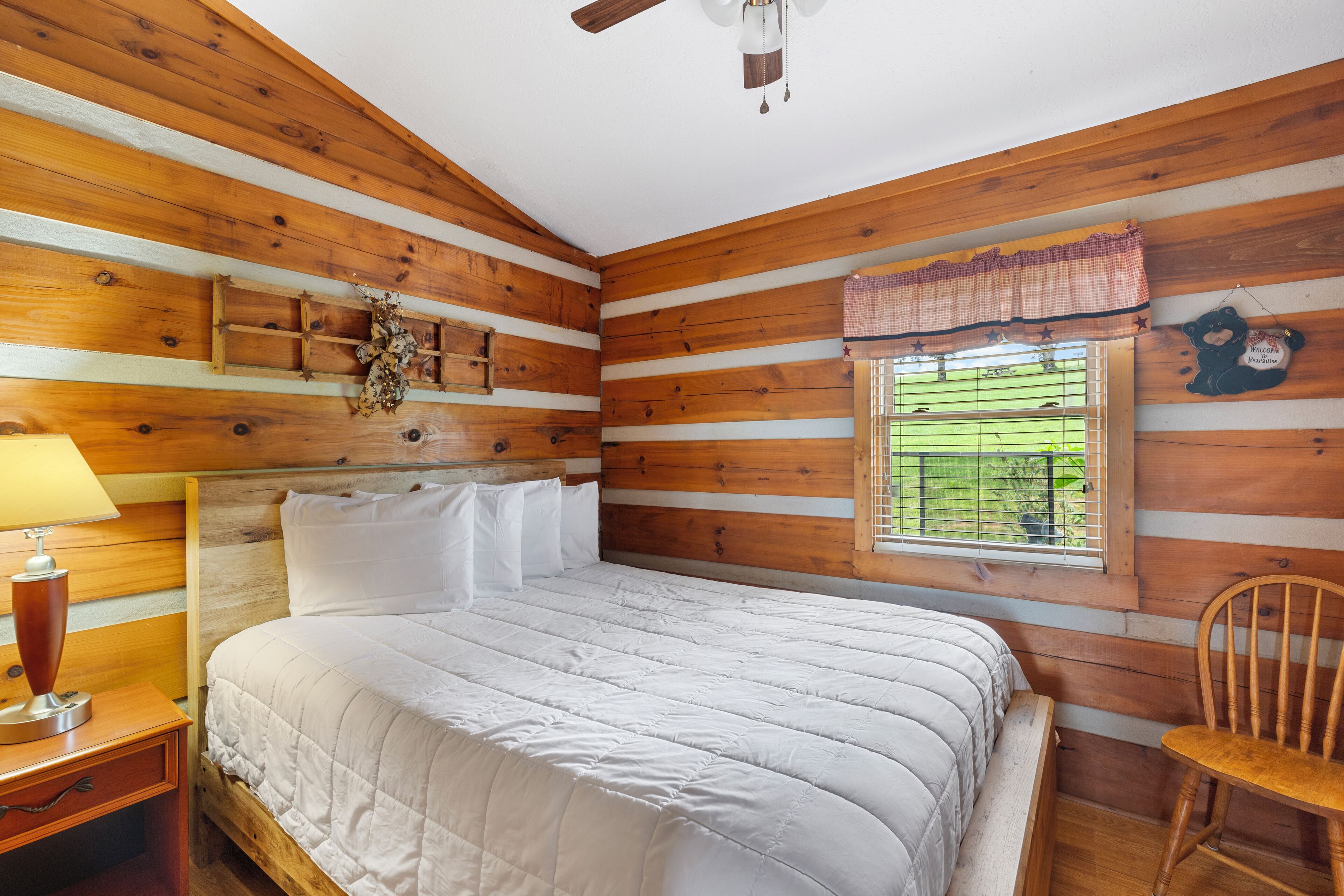 A wooden cabin-style bedroom with a bed, a nightstand, a lamp, a window with blinds, and a chair.