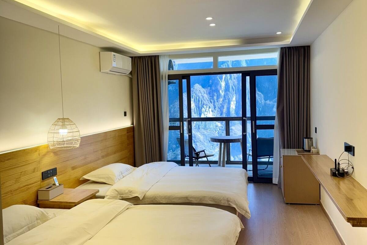 Een hotelkamer met twee bedden, een bureau, een stoel en uitzicht op bergen door grote ramen.