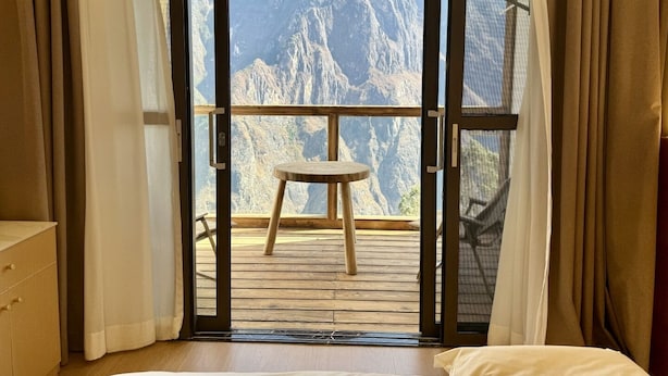 A room with a wooden floor, a bed, and a view of a mountainous landscape through a sliding glass door. - Tiger Leaping Half Way Guest House (Deqin)