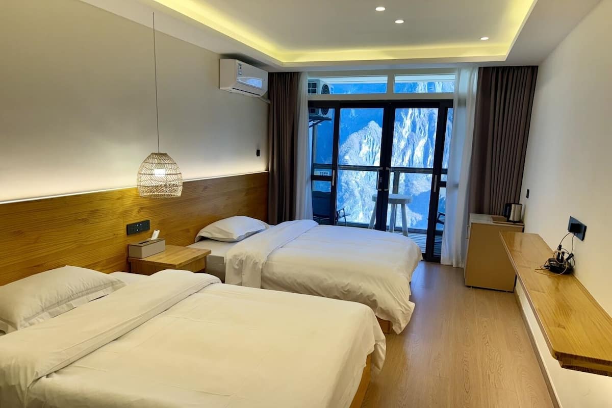 Hotelkamer met twee bedden, een houten hoofdbord, een nachtkastje met lamp, een raam met uitzicht op bergen en een bureau met stoel.