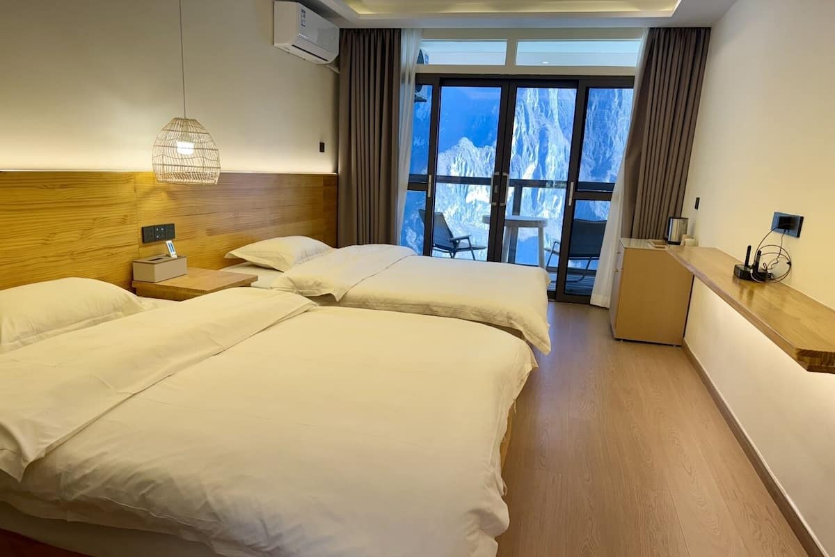Een hotelkamer met twee bedden, een houten hoofdbord, een bureau en uitzicht op bergen door grote ramen.