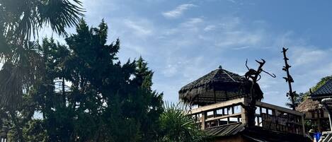 Une construction traditionnelle en bois, dotée d’un toit de chaume, entourée d’une végétation luxuriante et de grands arbres.