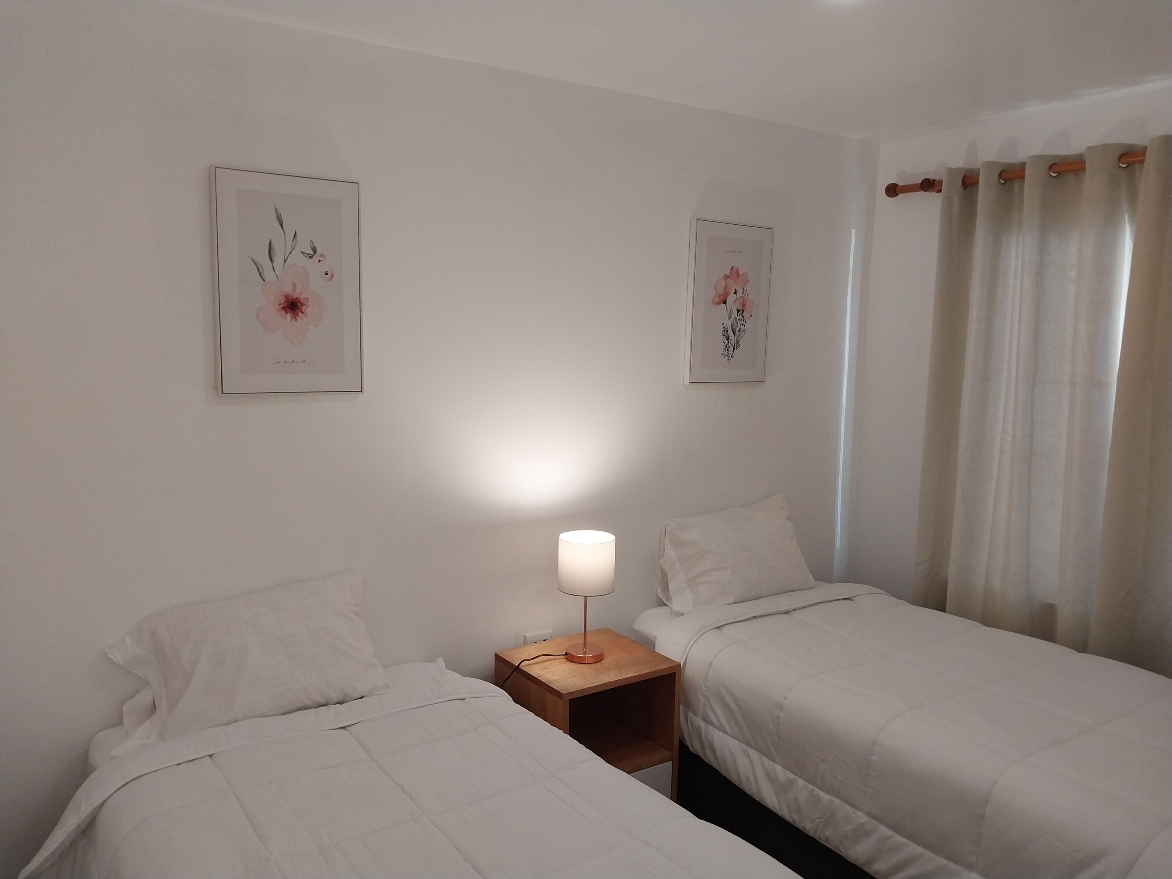 Et soveværelse med to senge, et natbord med lampe, og to rammede blomsterbilleder på væggen.