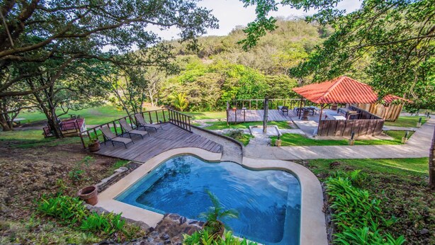 Un espace piscine doté d’une terrasse en bois, d’une petite piscine et d’un coin salon couvert avec un toit rouge.