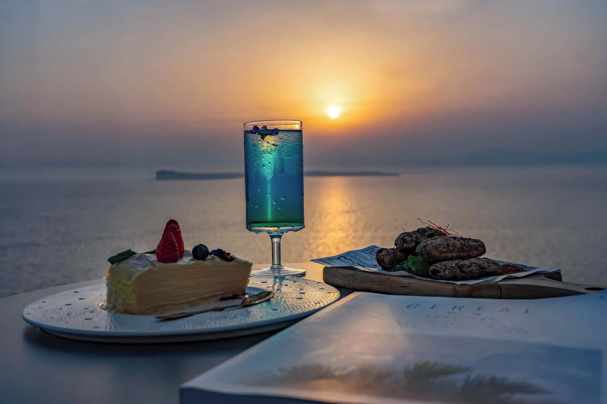 A dessert plate with a slice of cake and a glass of blue drink, a bowl of nuts, and a menu on a table by a body of water at sunset.