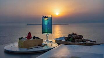 A dessert plate with a slice of cake and a glass of blue drink, a bowl of nuts, and a menu on a table by a body of water at sunset.