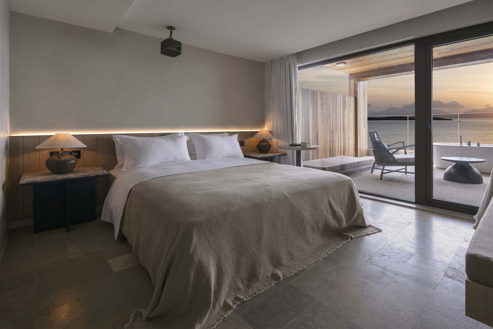 A modern hotel room with a large bed, bedside lamps, a small table, and a balcony with a view of the sea.