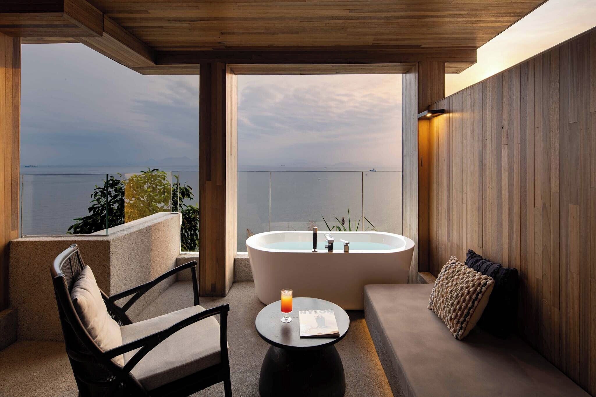 A modern bathroom with a freestanding bathtub, a sofa, and a chair, overlooking a body of water.