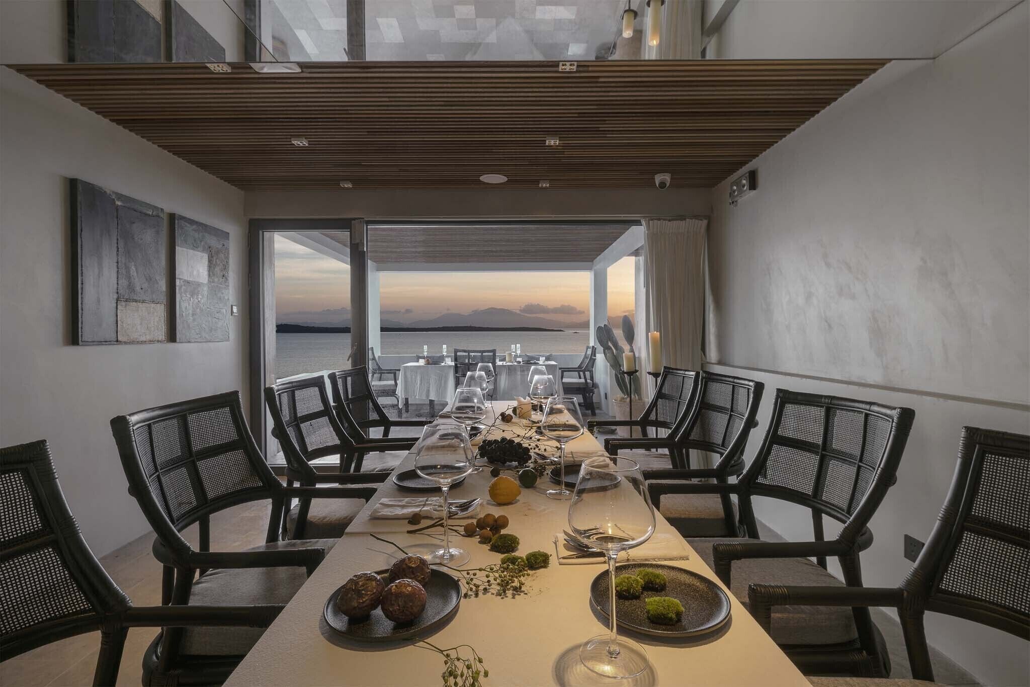 A dining area with a long table set for a meal, chairs on both sides, and a view of the sea through large windows.