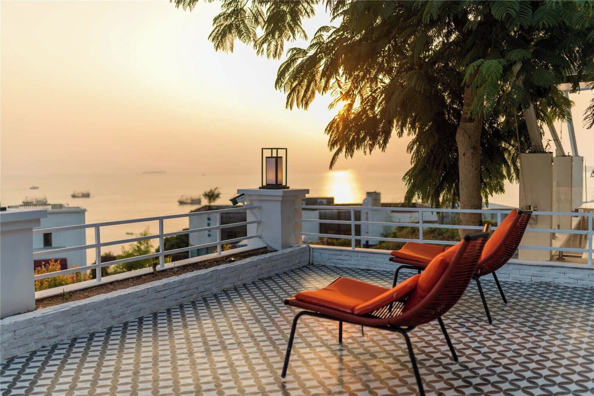 A rooftop terrace with a patterned floor, two orange chairs, a railing, and a view of the sea at sunset.
